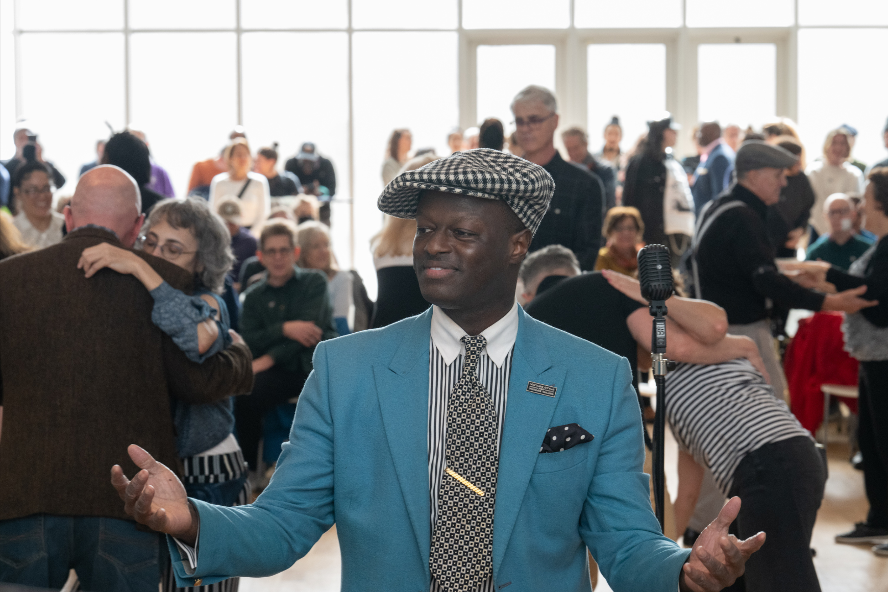 Performer Dandy Wellington dressed in a blue suit and patterned cap stands in front of a microphone, engaging with a lively crowd of people dancing and socializing in a bright, sunlit room